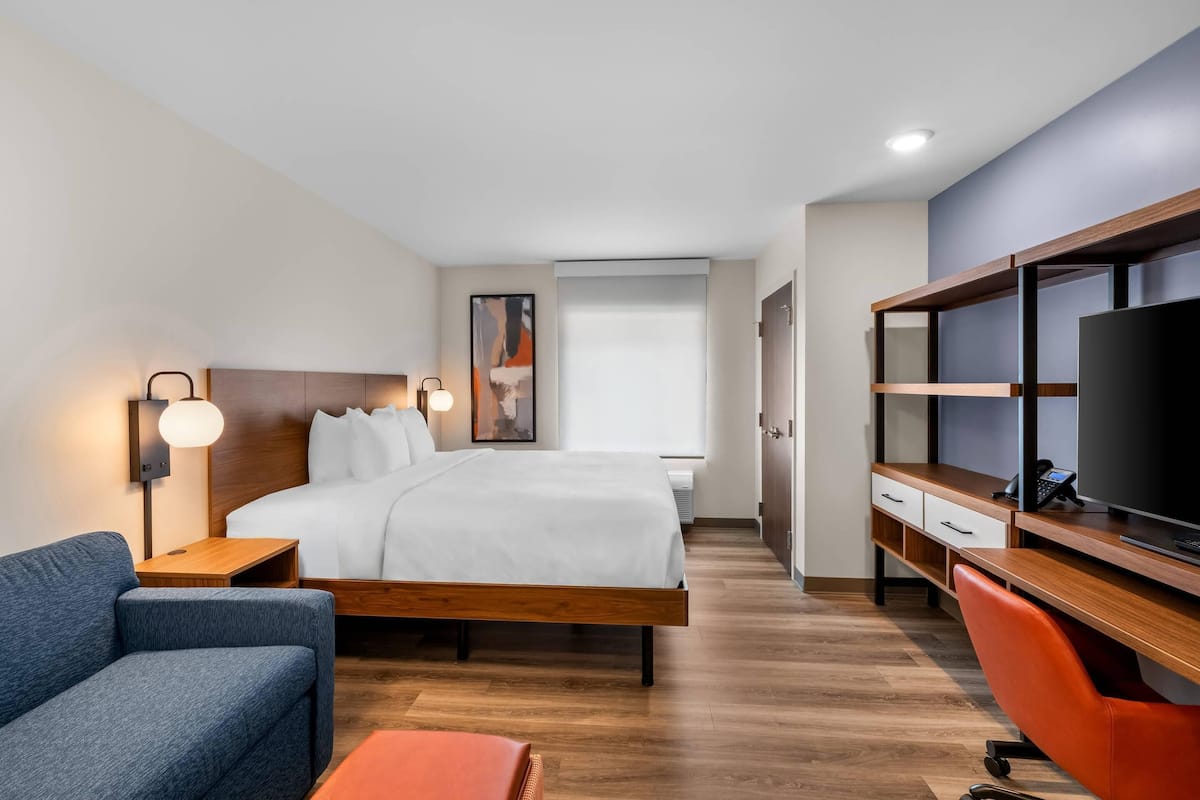 A hotel room with a bed, a desk with a TV, a chair, a sofa, and a nightstand with a lamp.