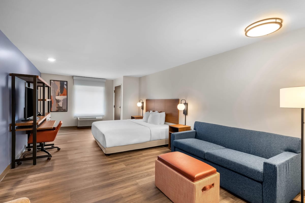 A modern hotel room with a bed, a blue sofa, a wooden desk, and a lamp.