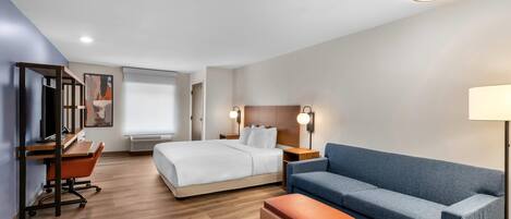 A modern hotel room with a bed, a blue sofa, a wooden desk, and a lamp.