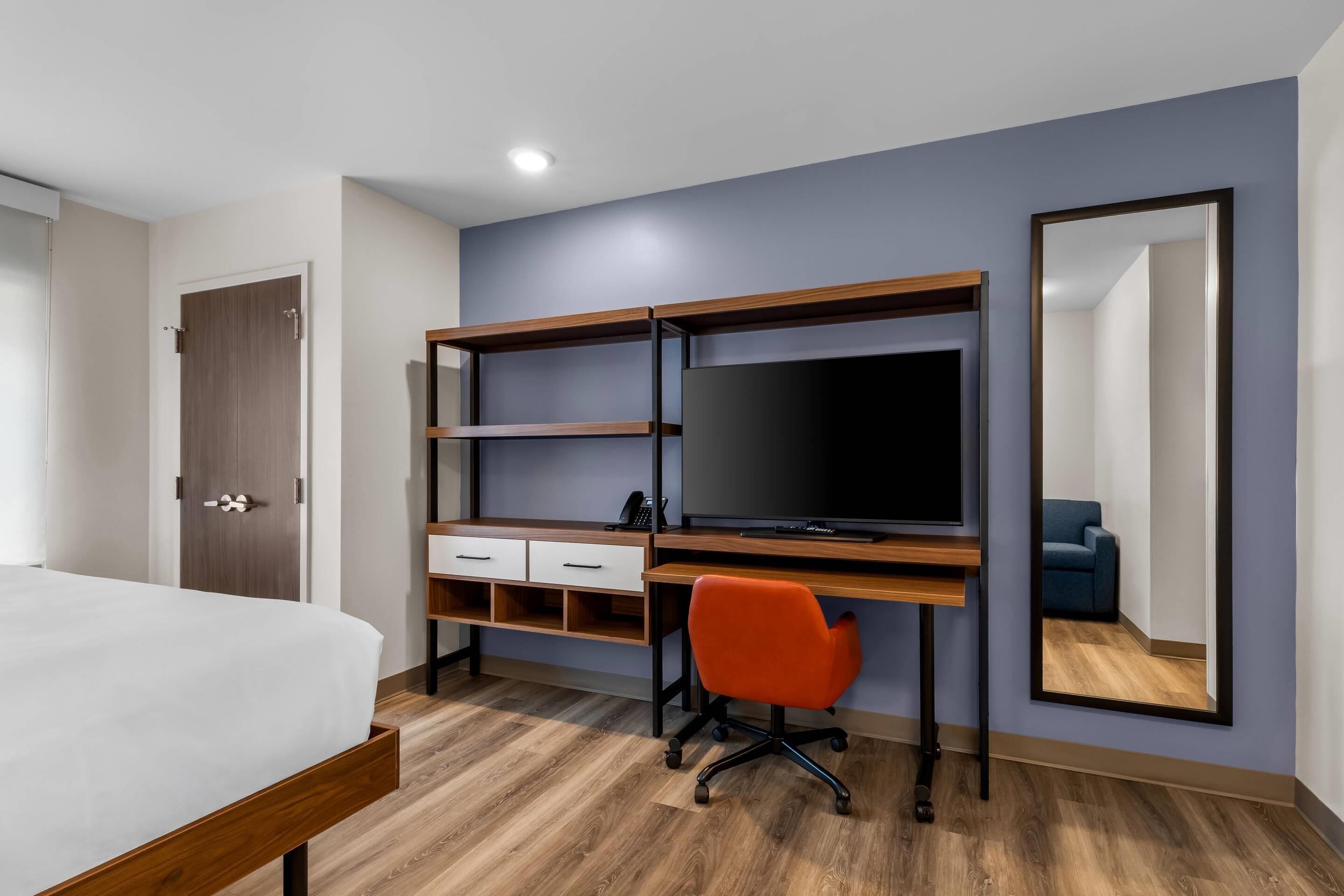 A hotel room with a bed, a desk with a TV, a chair, and a mirror.