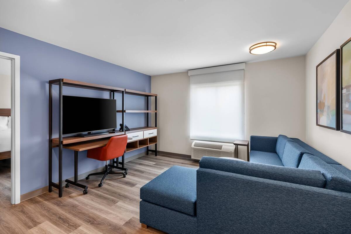 A modern living room with a flat-screen TV, a wooden desk, a red office chair, a blue sofa, and a large window with blinds.