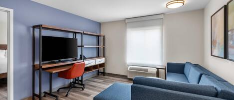 A modern living room with a flat-screen TV, a wooden desk, a red office chair, a blue sofa, and a large window with blinds.