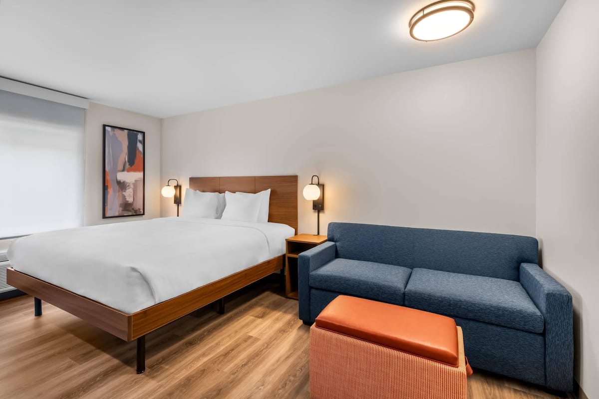 A hotel room with a bed, a blue sofa, a small orange ottoman, and two wall-mounted lamps.