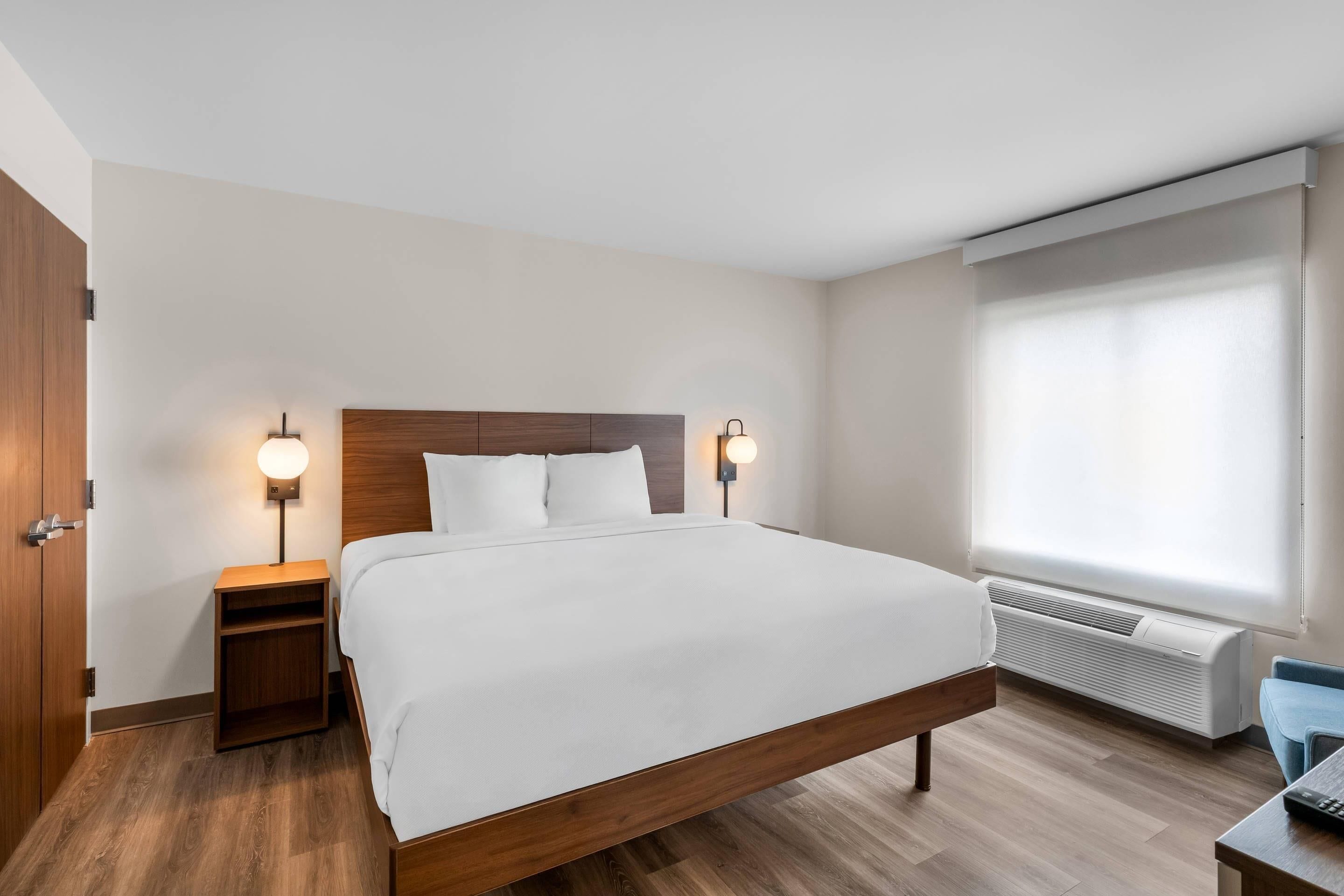 Een hotelkamer met een houten bed, nachtkastjes met lampen, een raam met zonwering en een wandairconditioner.