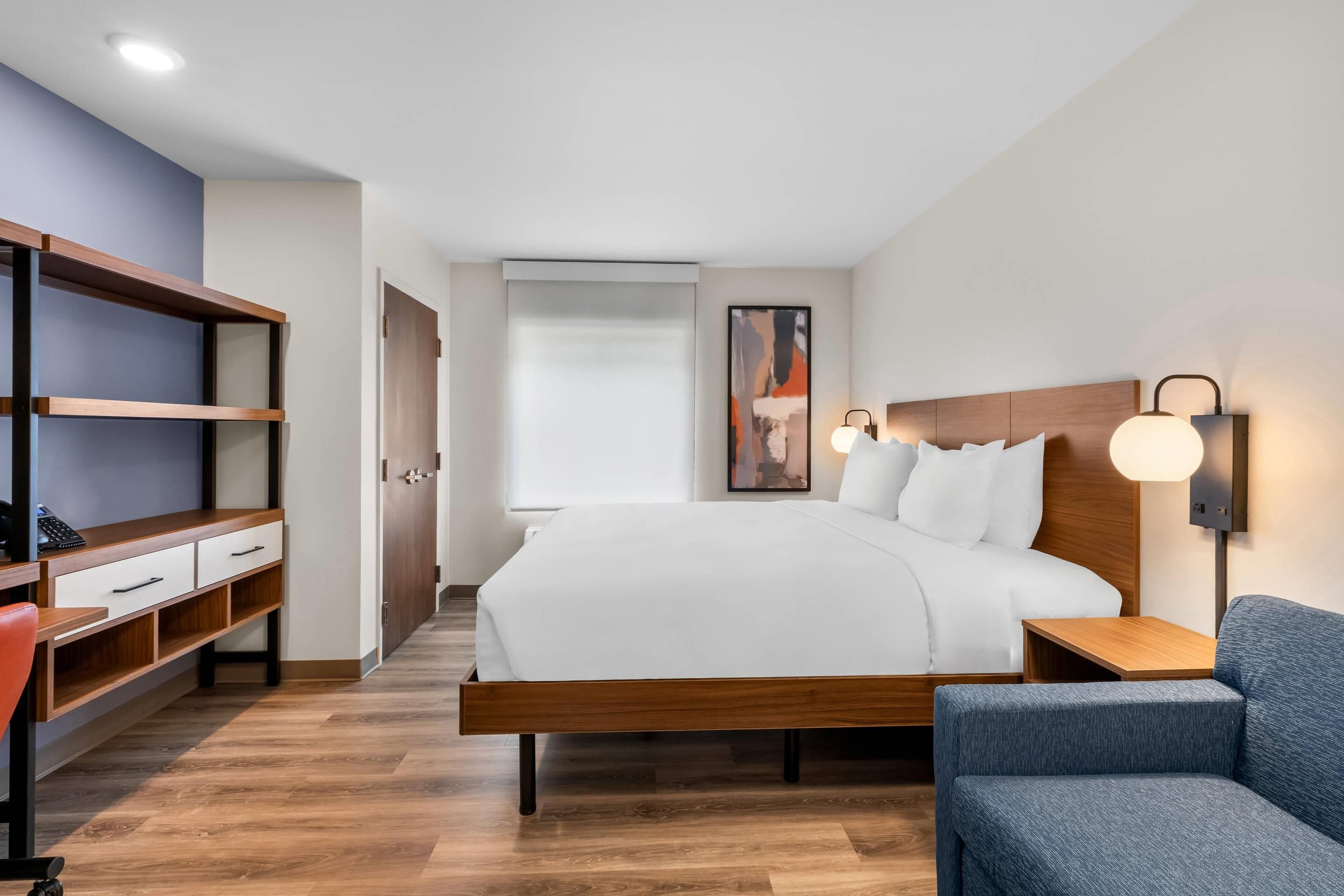 A modern hotel room with a wooden bed, a desk with a chair, a blue armchair, and a wall-mounted lamp.