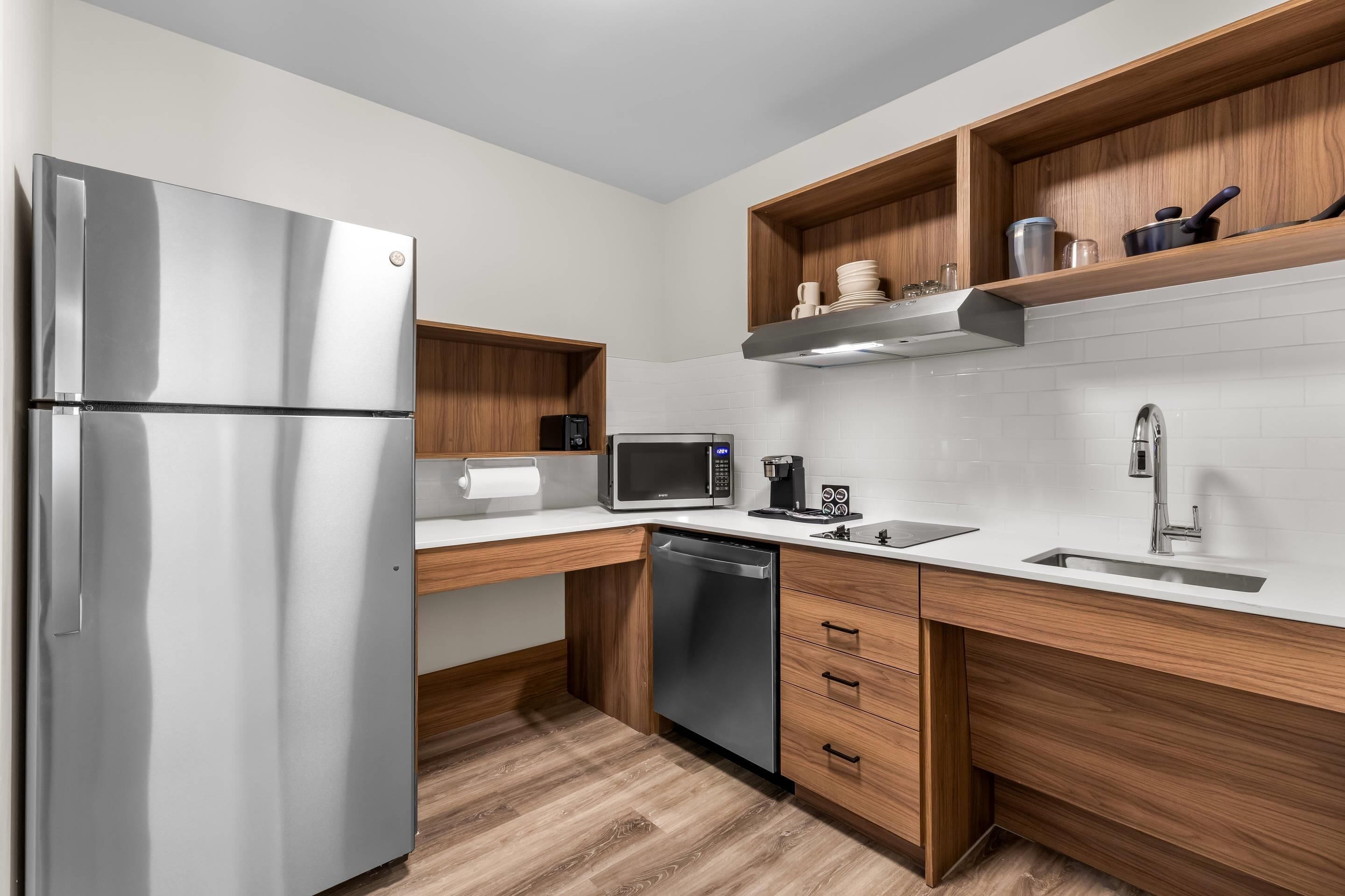 Una cocina moderna con refrigerador de acero inoxidable, microondas, lavavajillas, armarios de madera y salpicadero de azulejos blancos.