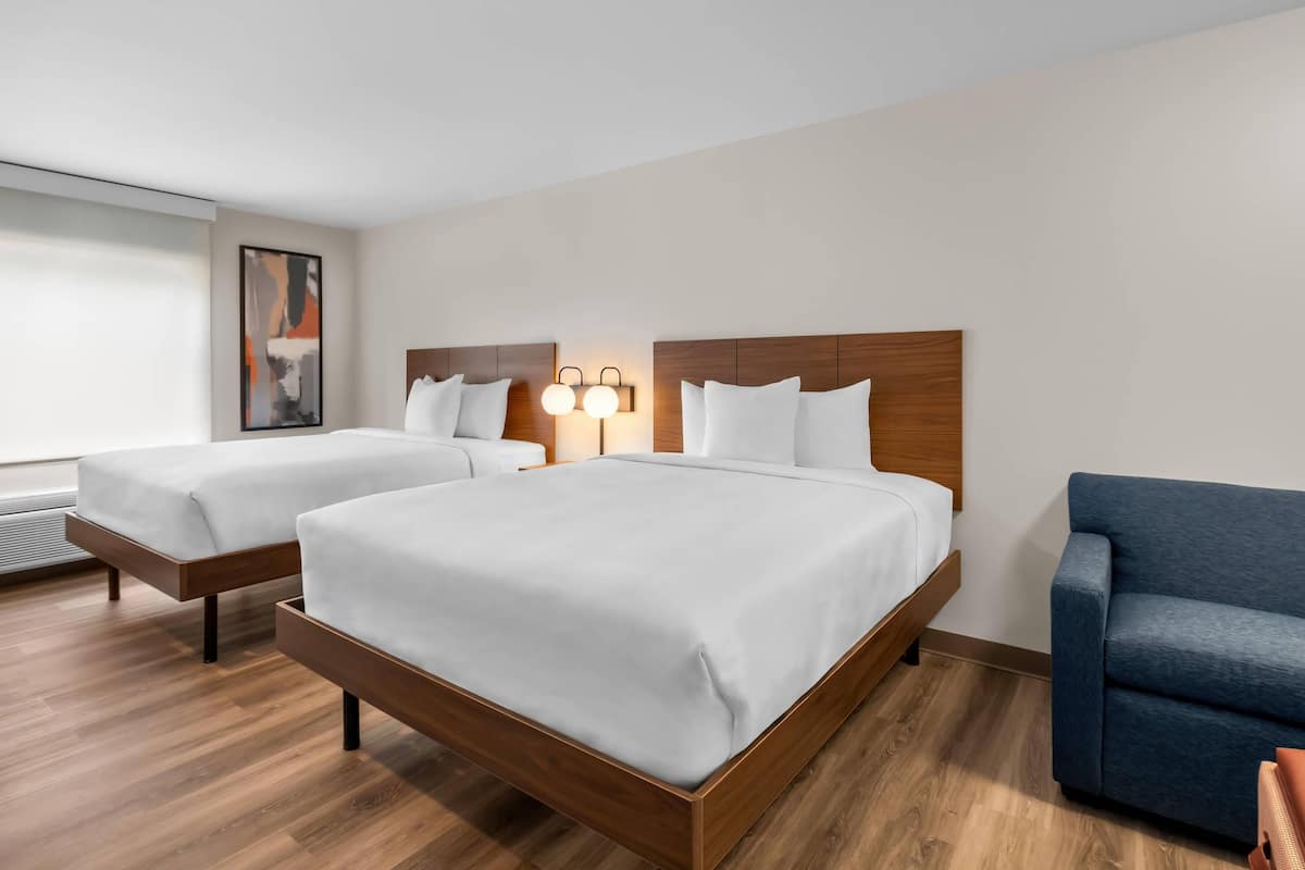 A hotel room with two beds, a blue armchair, and a wall-mounted light fixture.