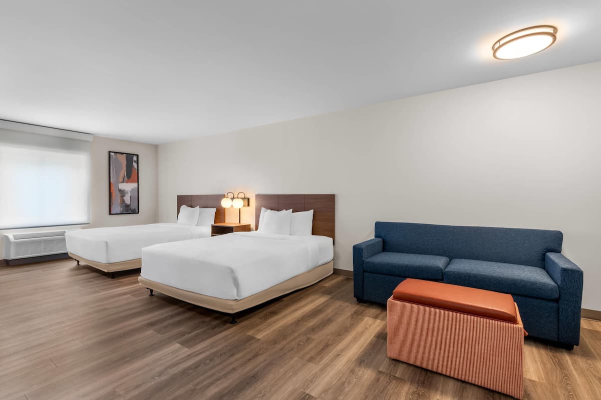 A hotel room with two beds, a sofa, an ottoman, a nightstand, and a wall-mounted light fixture.