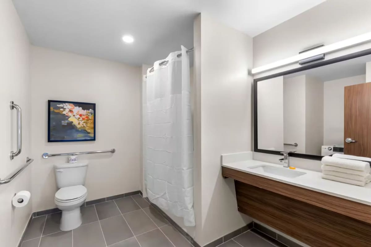 A bathroom with a toilet, a shower curtain, a sink with a mirror, and a handrail.