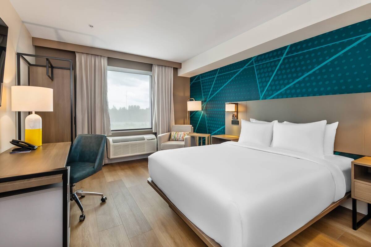 A hotel room with a large bed, a desk with a chair, a window with curtains, and a modern wall design.