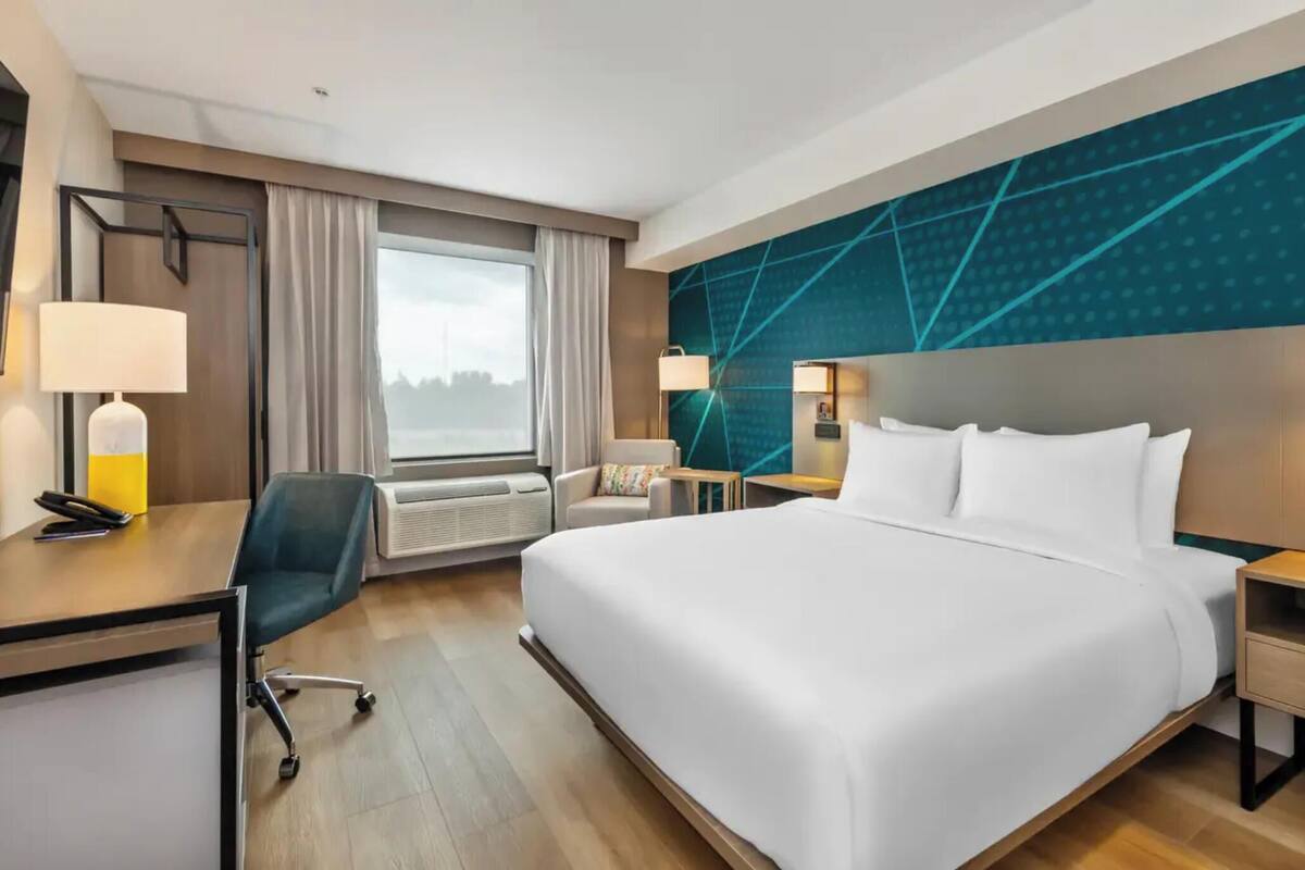 A hotel room with a large bed, a desk with a chair, a window with curtains, and a wall with geometric patterns.