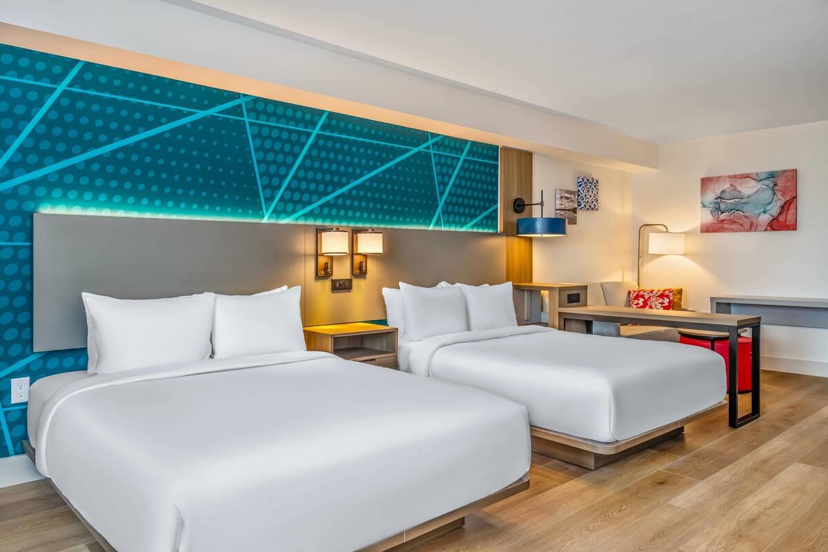 A hotel room with two beds, a desk, a chair, a nightstand, and a wall with geometric patterns and artwork.