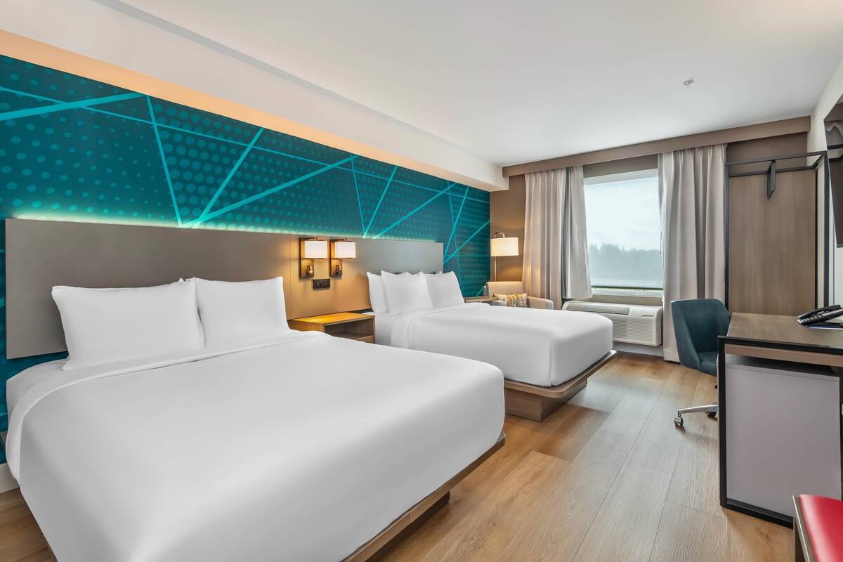 A hotel room with two beds, a desk, a chair, a desk lamp, and a view of the outdoors.