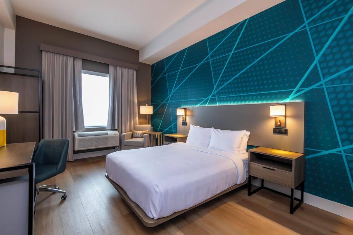 A modern hotel room with a large bed, a desk, and a chair. There is a window with curtains, a wall with a geometric pattern, and two bedside lamps.
