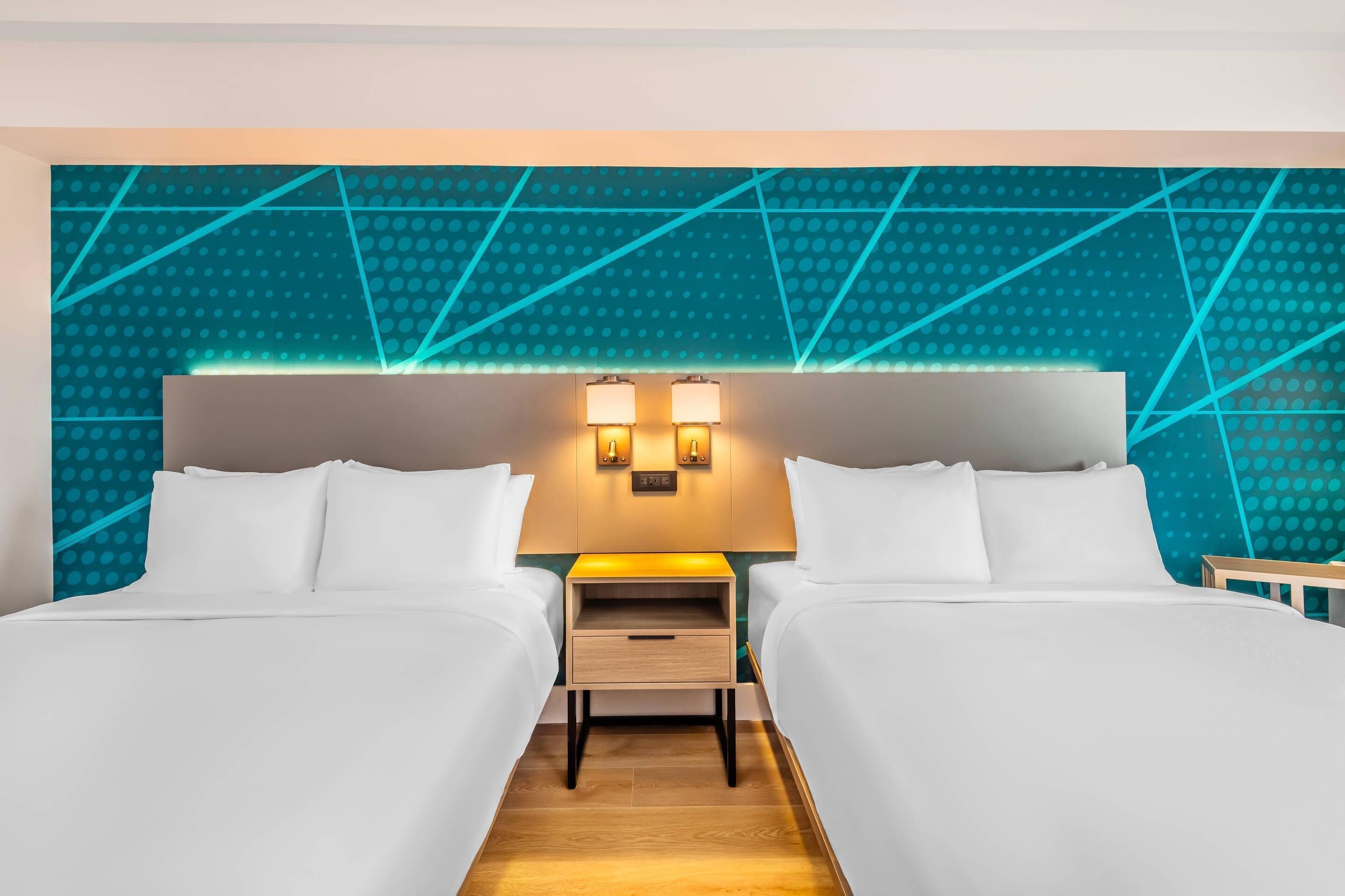 Una habitación de hotel con dos camas, una mesita de noche con lámpara y un papel tapiz de diseño geométrico.