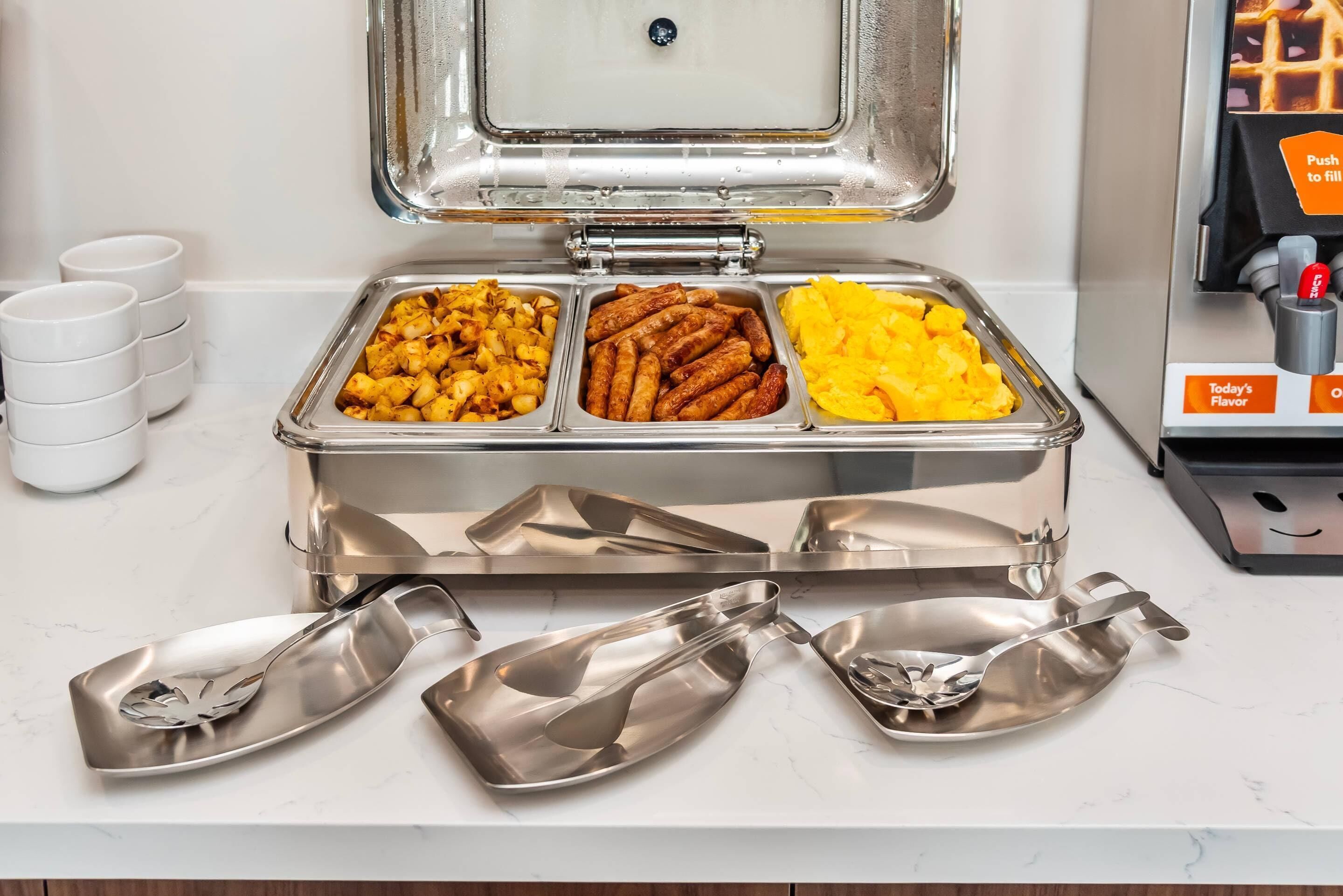 A buffet setup with three food containers: one with fried potatoes, one with sausages, and one with mashed potatoes.