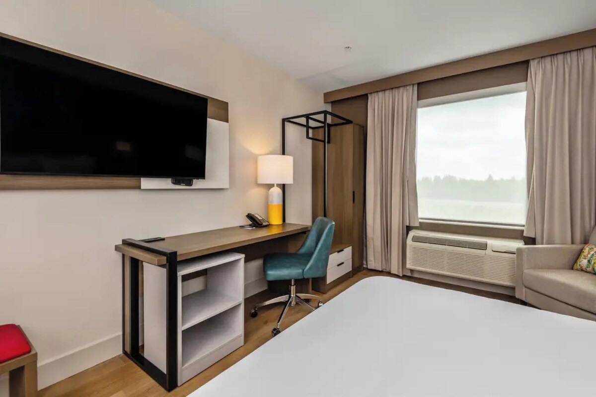 Une chambre d’hôtel équipée d’un lit, d’un bureau, d’une chaise, d’une télévision, d’une lampe et d’une grande fenêtre avec des rideaux.