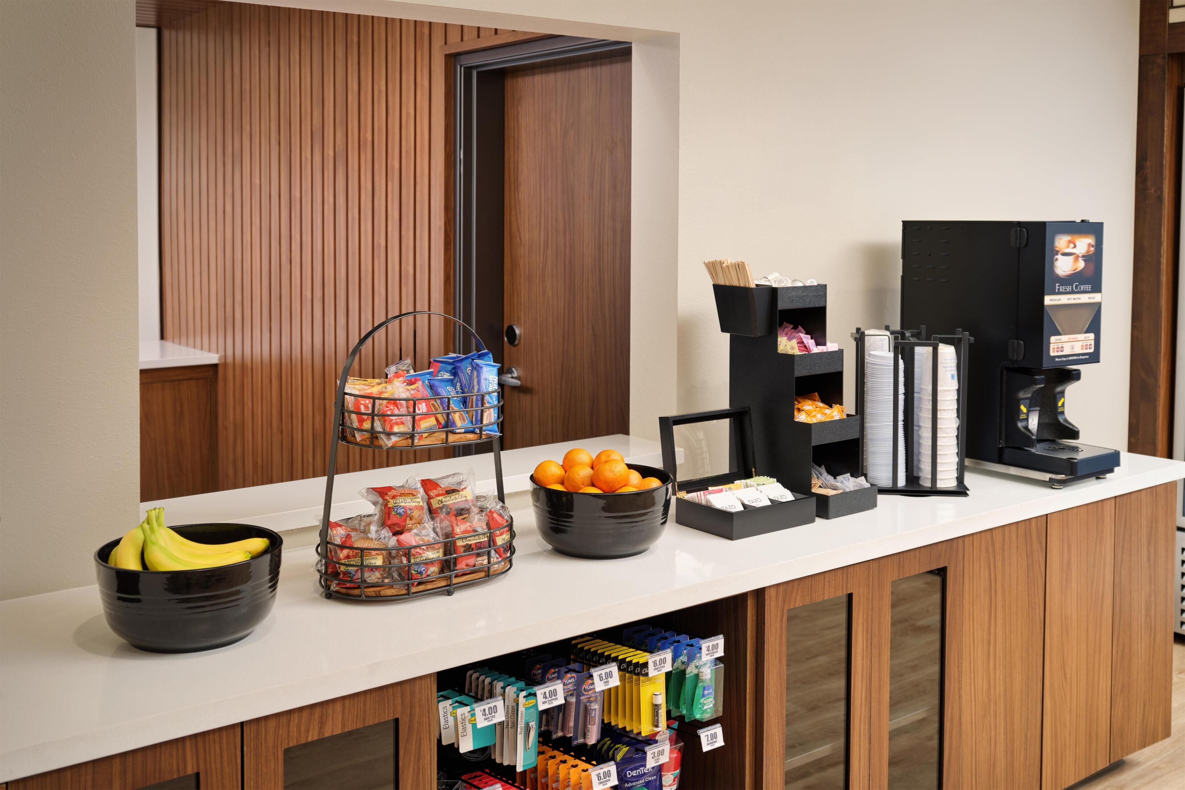 A hotel reception area with a counter, a coffee machine, a fruit bowl, a snack basket, and a beverage dispenser.