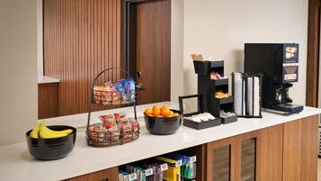 A hotel reception area with a counter, a coffee machine, a fruit bowl, a snack basket, and a beverage dispenser.