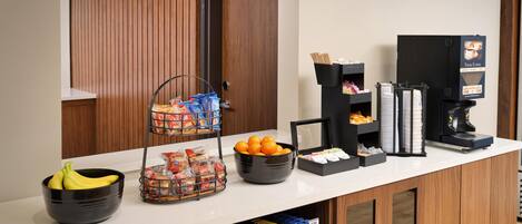 Ein Hotel-Empfangsbereich mit Theke, Kaffeeautomat, Obstschale, Snackkorb und Getränkespender.
