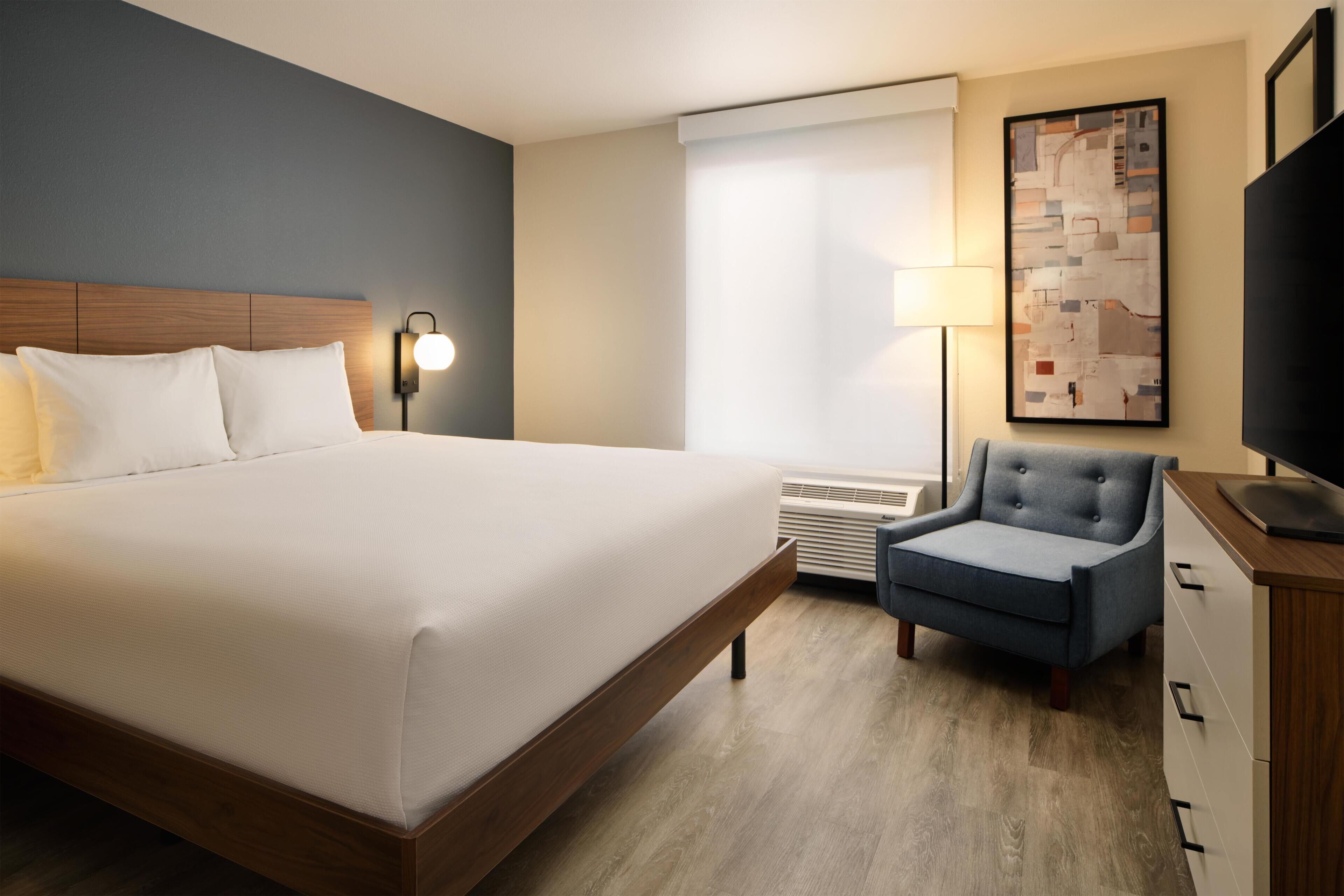 Une chambre d’hôtel avec un grand lit, une chaise, un bureau, une lampe, une télévision et une fenêtre avec des stores.