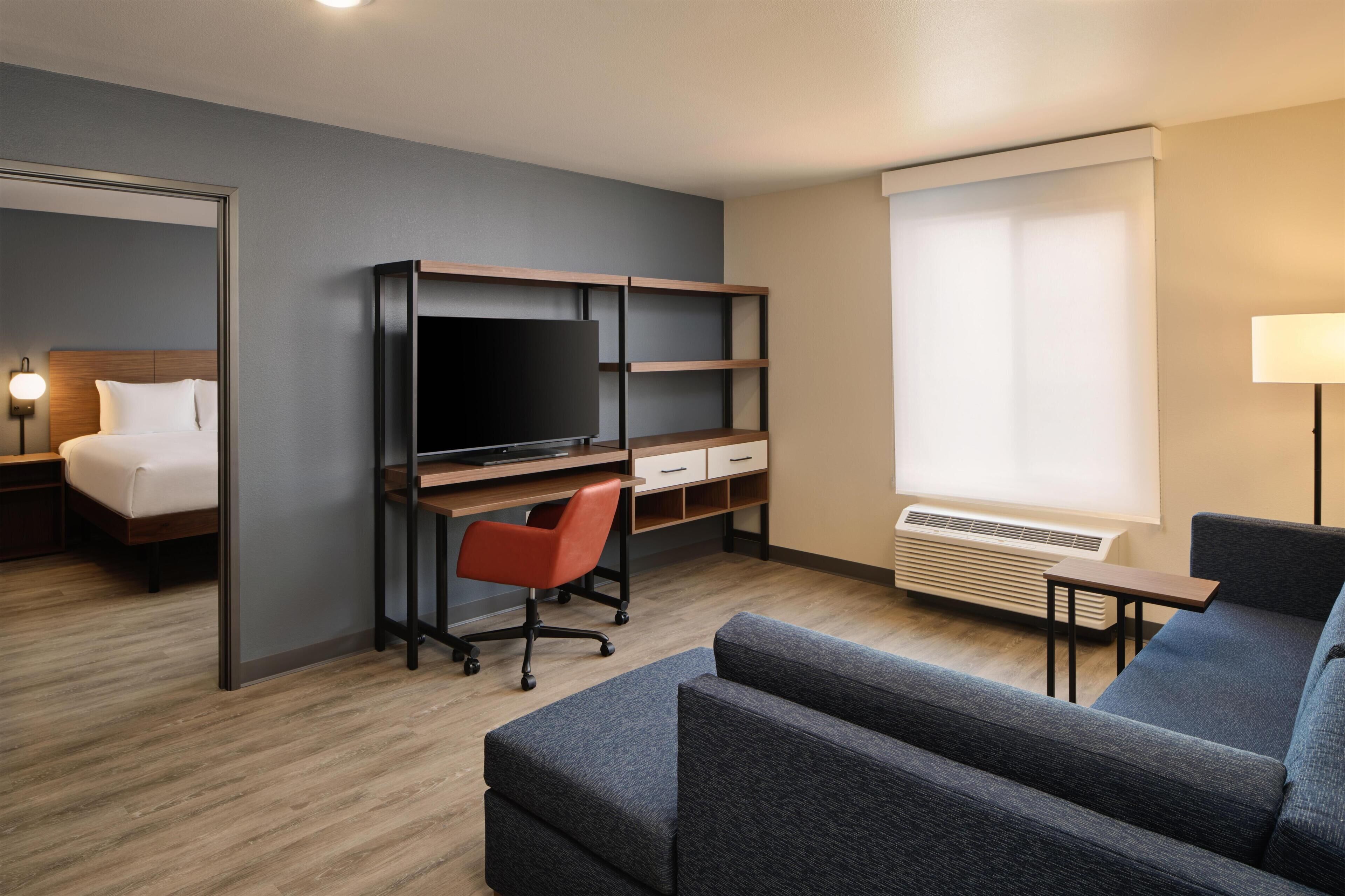 A hotel room with a bed, a desk with a TV, a sofa, and a window with blinds.
