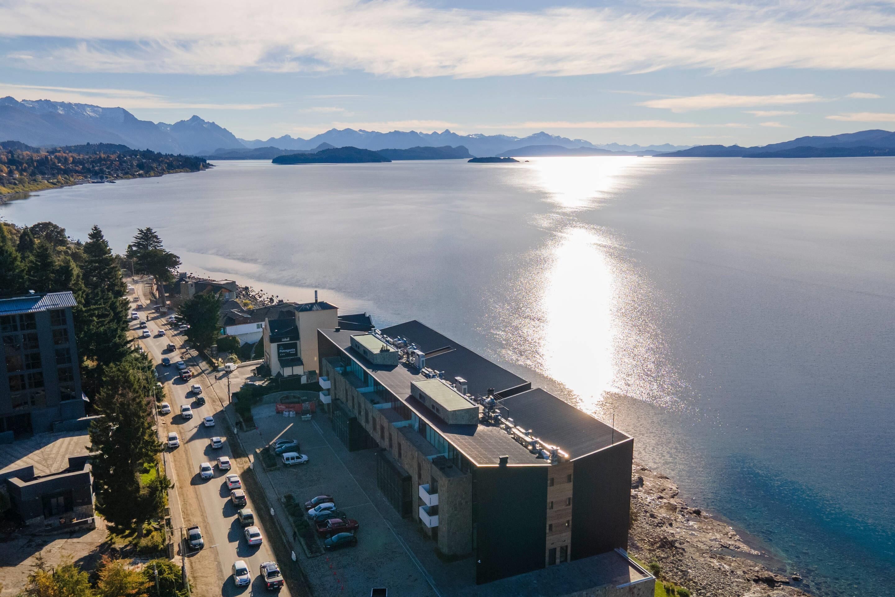 Une vue aérienne d’une zone côtière avec une vaste étendue d’eau, des montagnes en arrière-plan et un parking bordé de plusieurs bâtiments situés à proximité de l’eau.