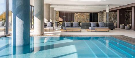 A modern indoor pool area with a clear pool, wooden decking, and seating areas with sofas and chairs.
