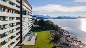 Un hotel moderno con varios balcones, piscina y vistas a un lago y montañas.
