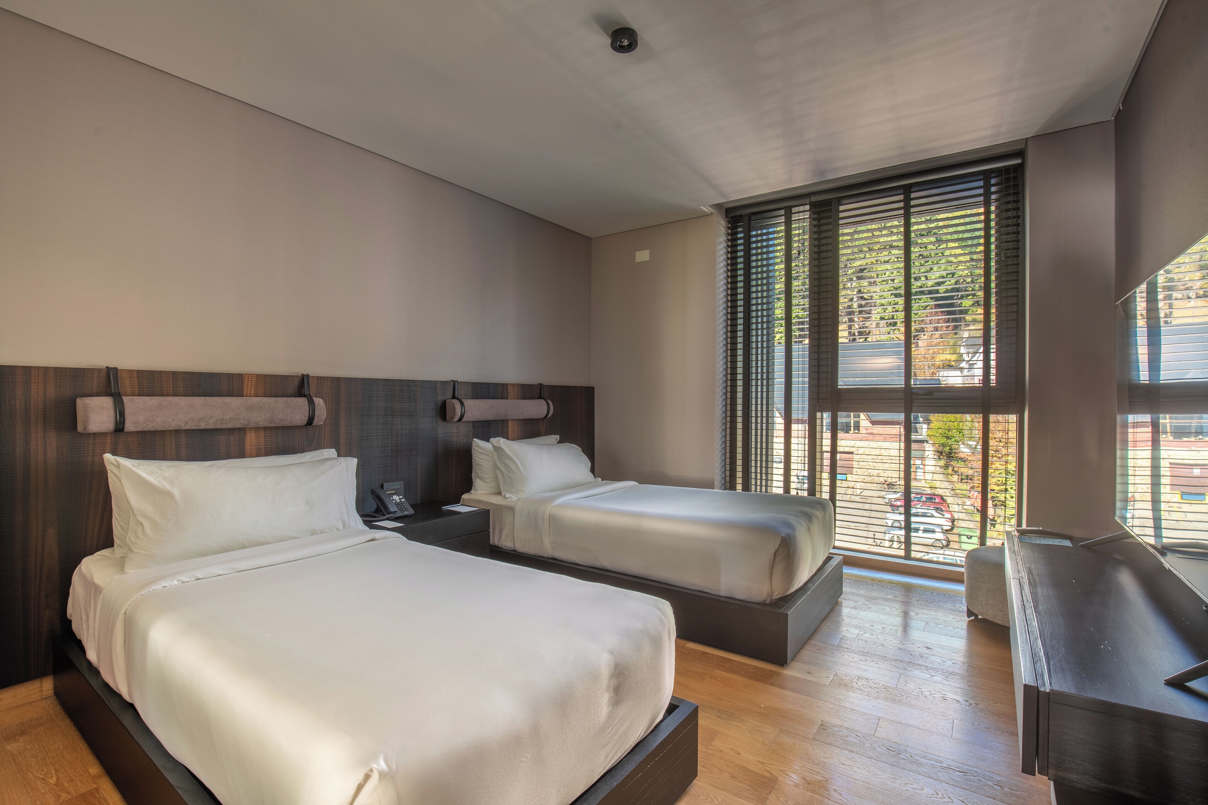 Moderni hotellihuone, jossa on kaksi sänkyä, työpöytä ja suuri ikkuna, jossa on sälekaihtimet.