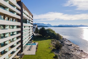 Ein modernes Hotel mit mehreren Balkonen, einem Poolbereich und Blick auf einen See und Berge.