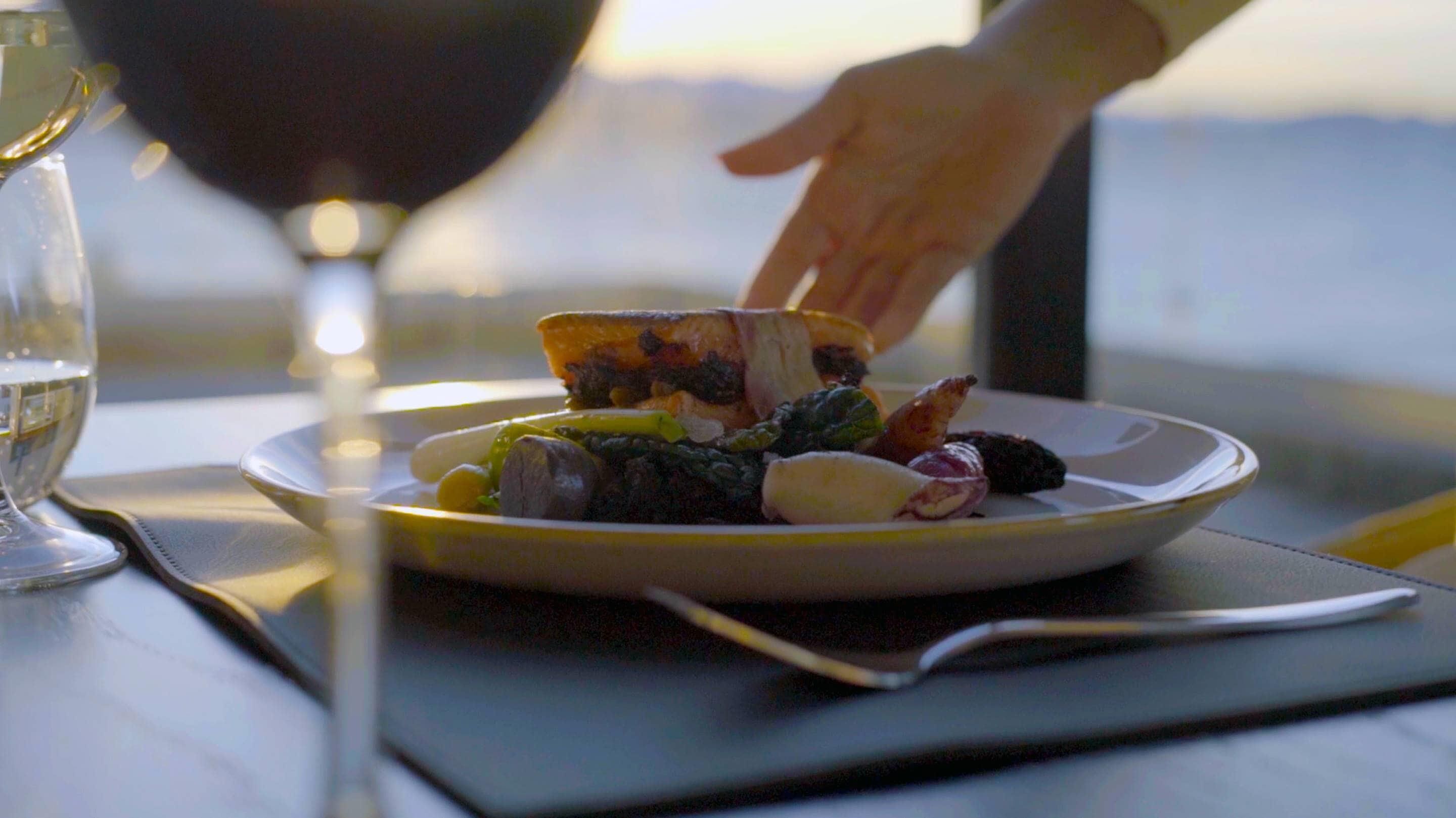 Un plato de comida con una mano acercándose, una copa de vino y vistas al exterior.