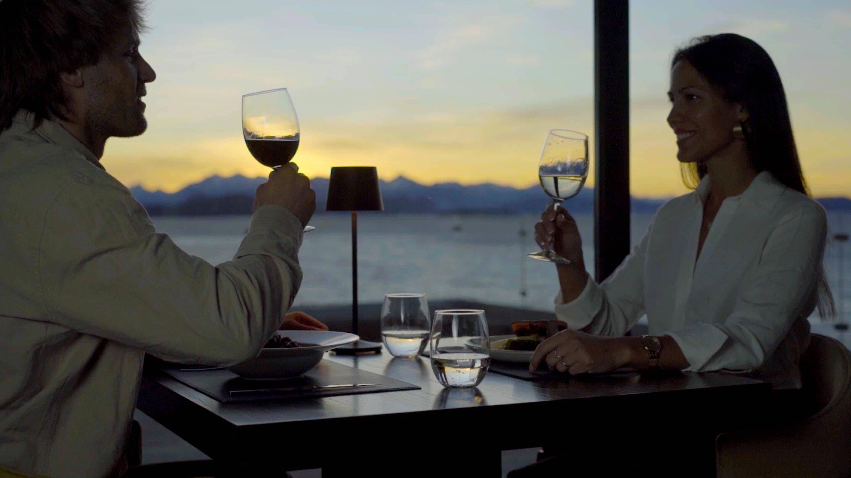 Um casal jantando à mesa com vista para o mar e montanhas ao fundo.