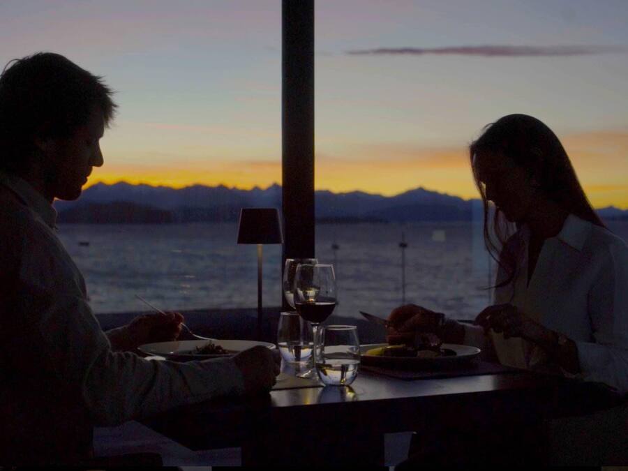 Um casal jantando à mesa com vista para o mar e montanhas durante o pôr do sol.