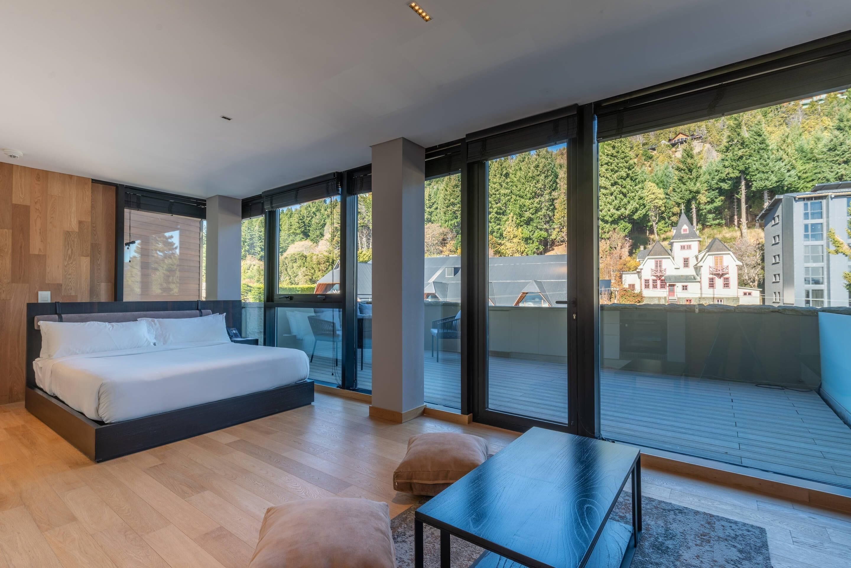 Un dormitorio moderno con una cama grande, una mesita pequeña y un balcón con vistas a casas y árboles.