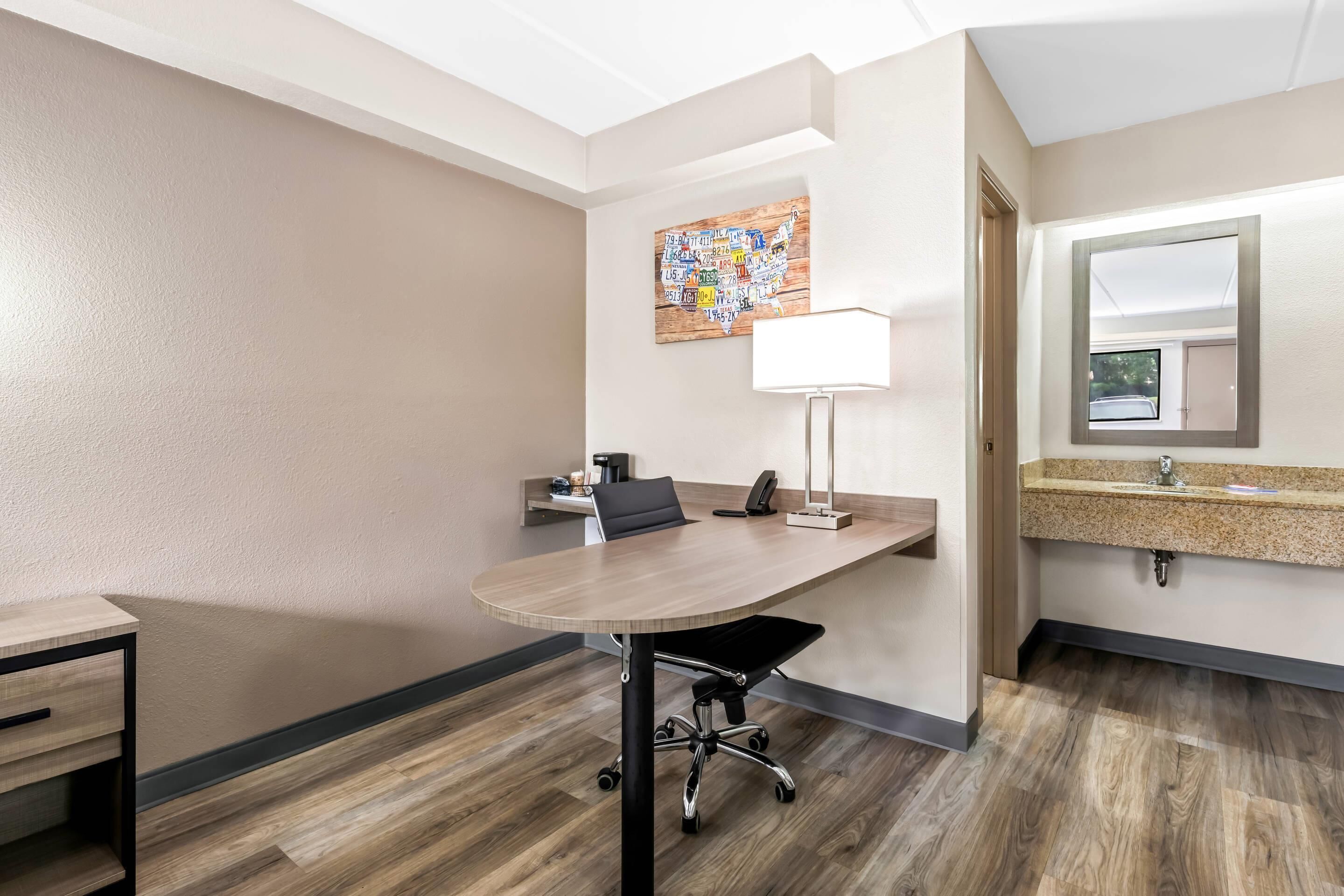 A hotel room with a wooden floor, a desk with a chair, a lamp, a mirror, and a wall art piece.
