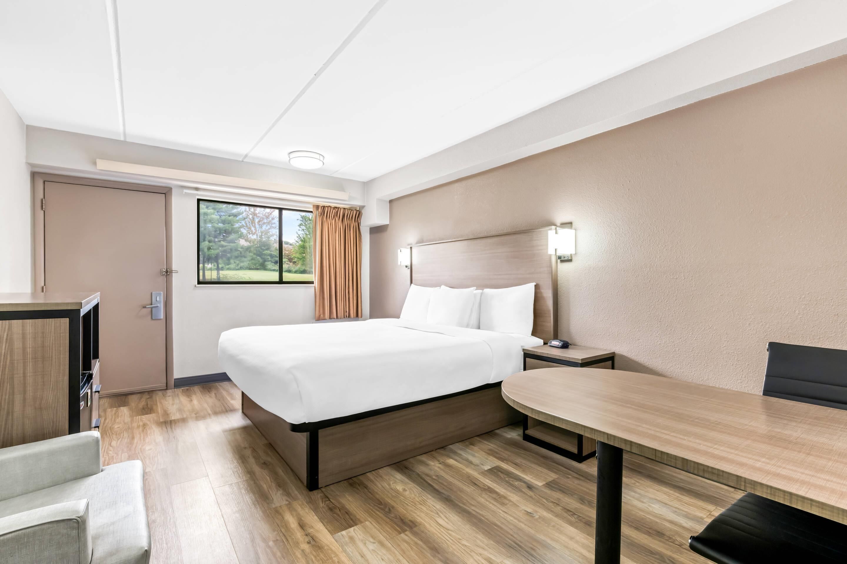 A hotel room with a bed, bedside table, lamp, and a view of the outdoors.