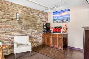 Una habitación con pared de ladrillo, una silla blanca, una estación de café con varias cafeteras y un cuadro de un paisaje.