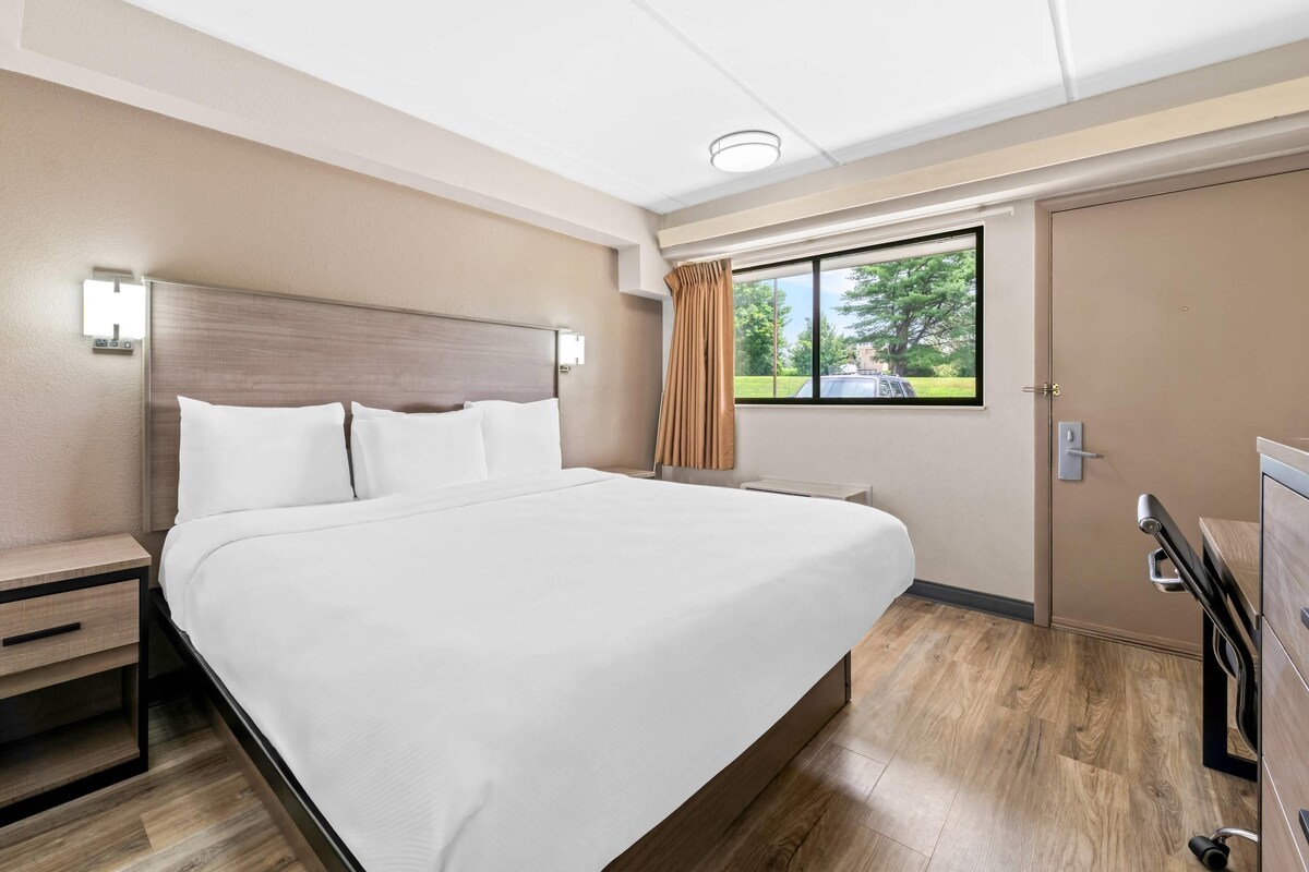 A hotel room with a large bed, a nightstand, a dresser, a window with curtains, and a view of the outdoors.