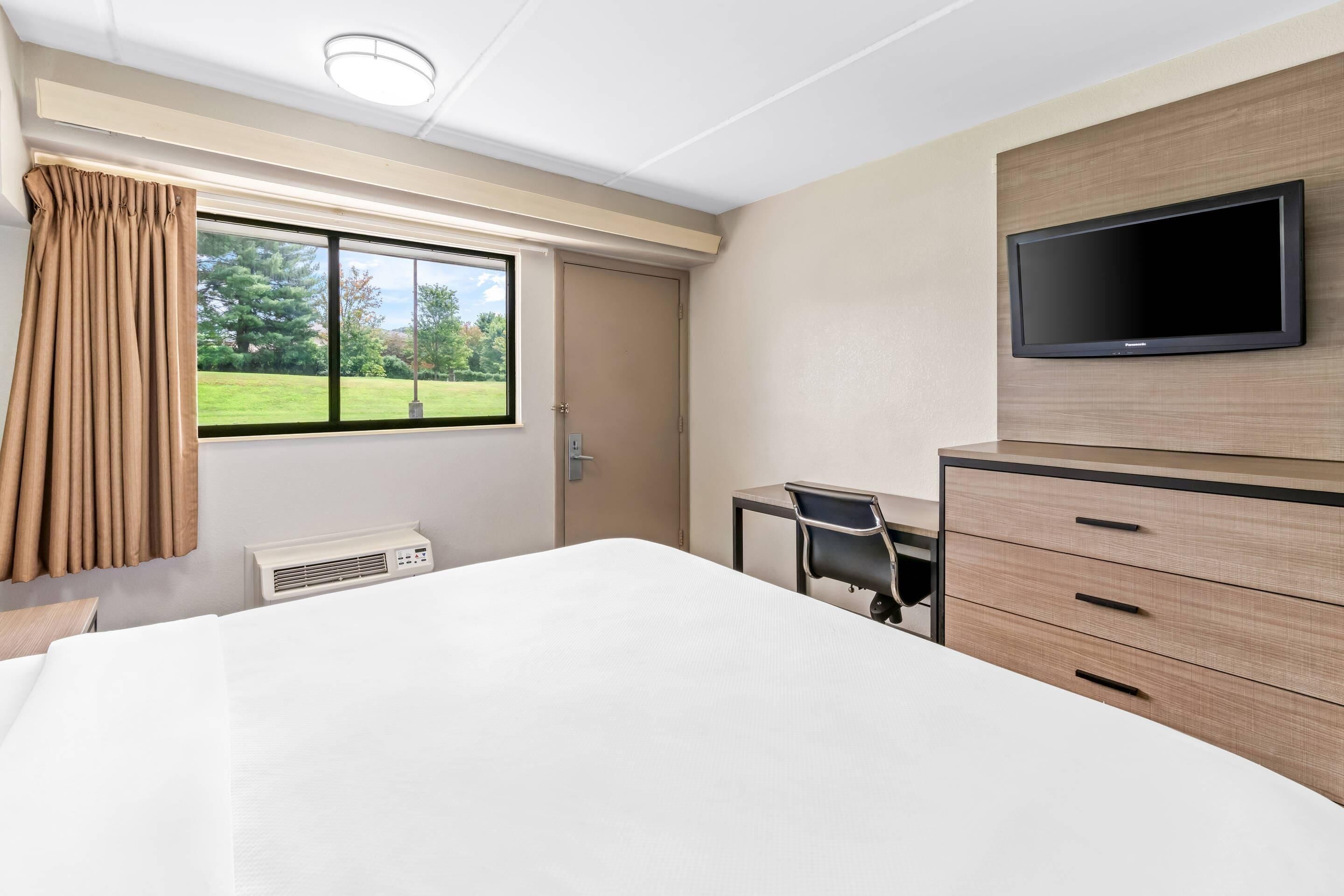 Habitación de hotel con cama, escritorio, silla, televisor y ventana con cortinas.