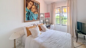 A bedroom with a large bed, a painting on the wall, a desk with a lamp, and a TV.
