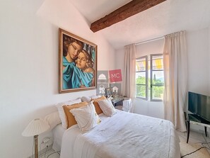A bedroom with a large bed, a painting on the wall, a desk with a lamp, and a TV.