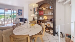 A dining area with a round table, chairs, and a chandelier. There is a view of the outdoors through large windows.
