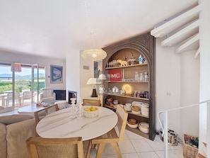 A dining area with a round table, chairs, and a chandelier. There is a view of the outdoors through large windows.