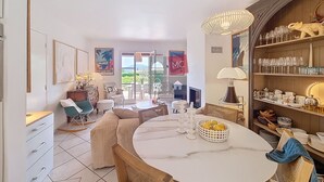 A spacious living area with a round marble table, a bookshelf filled with books and decorative items, and a view of the outdoors through large glass doors.