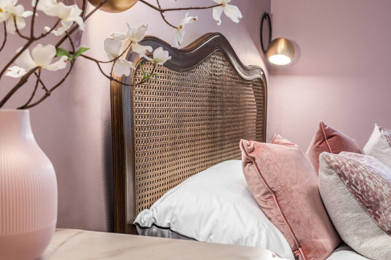 A bedroom with a woven headboard, white and pink pillows, and a vase with flowers.