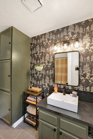 Une salle de bain avec un lavabo blanc, des meubles verts, un miroir avec éclairage intégré et un papier peint à motifs.