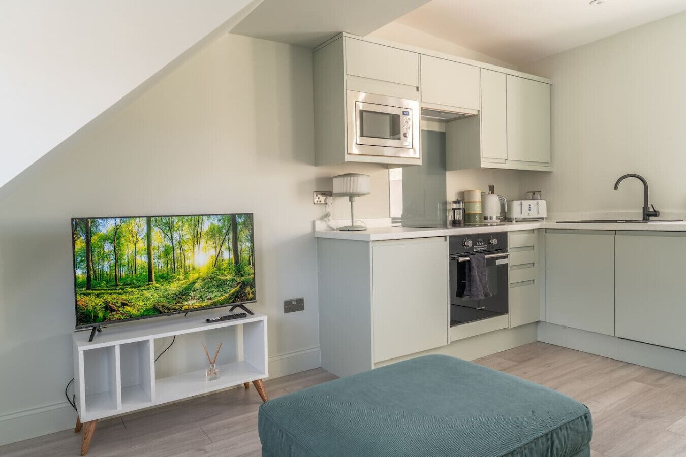 Ett modernt kök med inbyggd mikrovågsugn, ugn och diskho. Det finns en tv på ett vitt tv-bord med en bild av en skog, och en turkosfärgad pall i förgrunden.