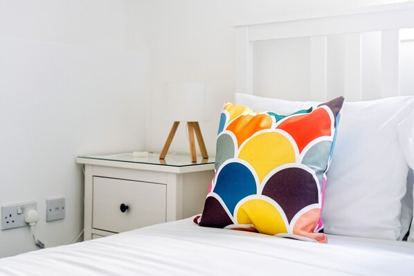 A bed with a colorful pillow, a white bedside table with a lamp, and a white wall with an electrical outlet. - Argyle Street - Apartment 3 (Glasgow)
