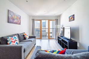 A modern living room with a grey sofa, colorful pillows, a flat-screen TV, and a large window with a city view. - Argyle Street - Apartment 3 (Glasgow)